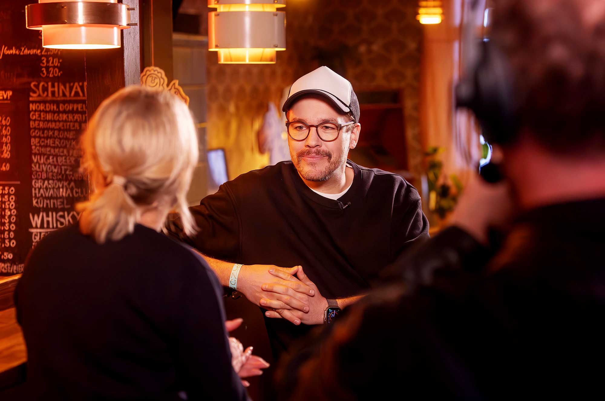 Jan van Weyde mit Cap und Brille im Interview vor einer Bar mit Getränkekarte